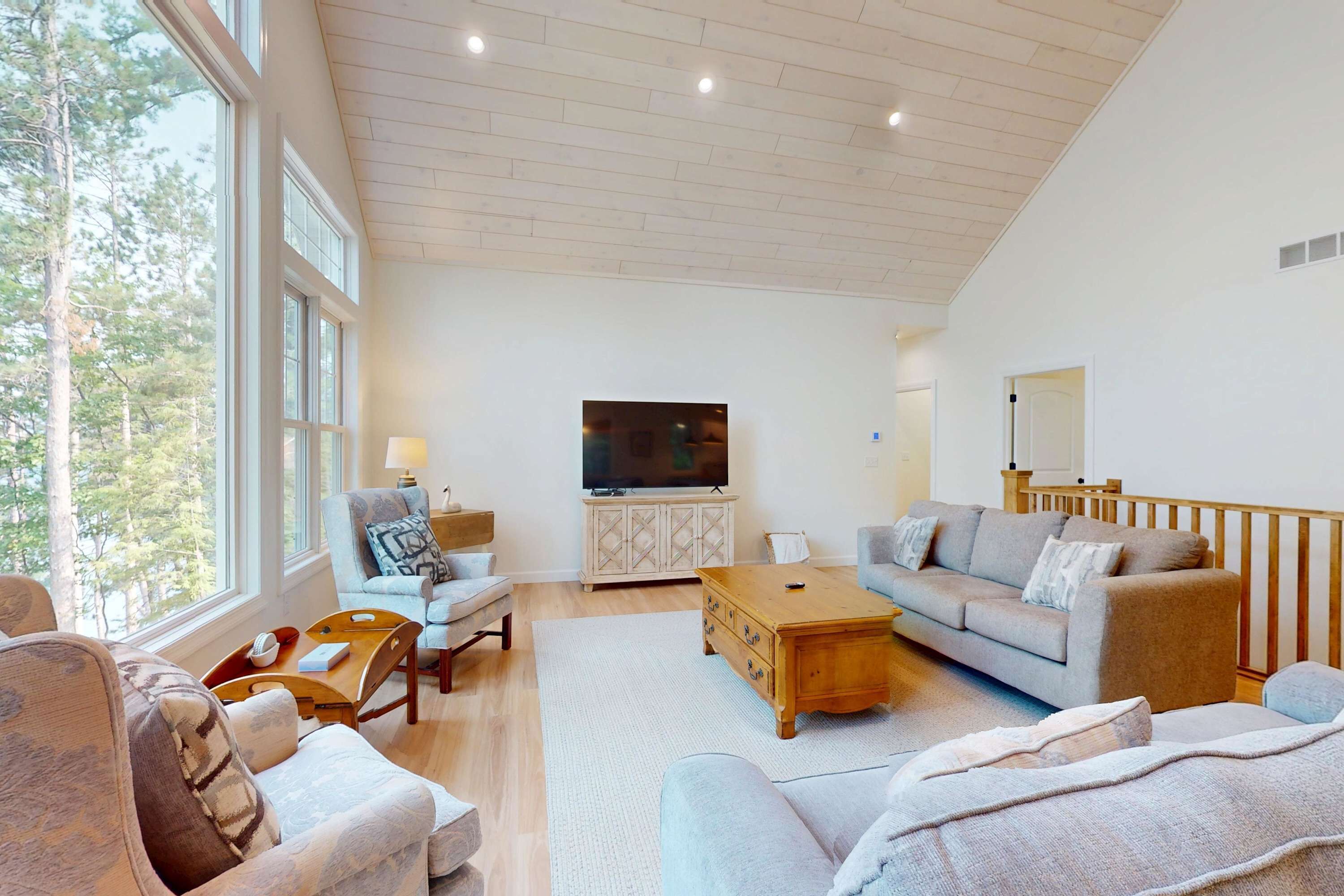 A spacious living room with a high ceiling, large windows, a flat-screen TV, and a cozy seating area with a sofa and armchairs.