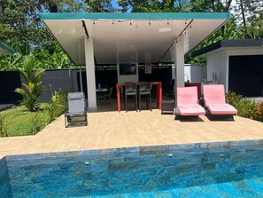 Ett område vid poolen med en överbyggd sittplats, solstolar och en pool.
