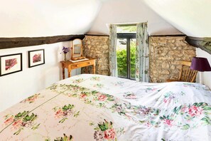 Ein Schlafzimmer mit schräg versiegelter Decke, Steinwänden, einem hölzernen Bettgestell, einem Nachttisch und einem Fenster mit Vorhängen.