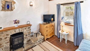 A cozy room with a stone fireplace, a television, a small table with a lamp, and a window with checkered curtains. - Holiday Cottage Near Axminster (Axminster)
