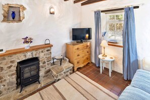 Ein gemütliches Zimmer mit Kamin aus Stein, einem Fernseher, einem kleinen Tisch mit Lampe und einem Fenster mit karierten Vorhängen.