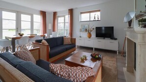 Ett modernt vardagsrum med en soffa, ett soffbord och en tv. Det finns fönster med gardiner, en öppen spis och en matplats med stolar.