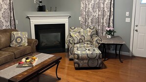 A living room with a fireplace, a ceiling fan, a sofa, a patterned armchair, and a coffee table with fruit. - CORP HOUSING:::
Spacious 5-bedroom house in amazing Winston-Salem with AC (Winston-Salem)