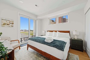 Ein modernes Schlafzimmer mit einem großen Bett, einem Nachttisch, einem Fenster mit Blick auf Gebäude und einem Balkon.
