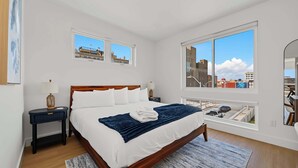 A bedroom with a large bed, two bedside tables, a window with a city view, and a mirror on the wall. - Coastal Loop Retreat | Rooftop Deck + Garage (Atlantic City)