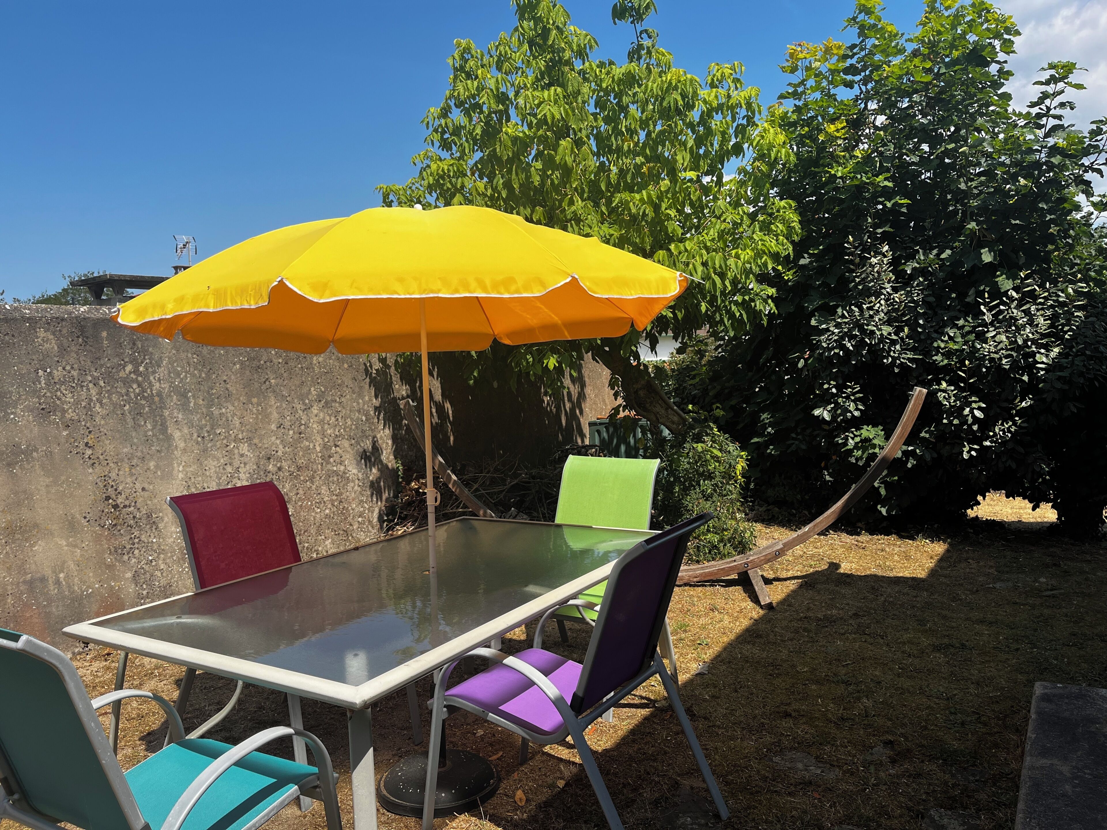 An outdoor dining area with a table and chairs, a yellow umbrella, and a garden backdrop.