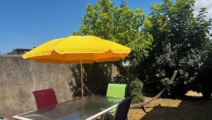 Un área de comedor al aire libre con mesa y sillas, un paraguas amarillo y un jardín de fondo.