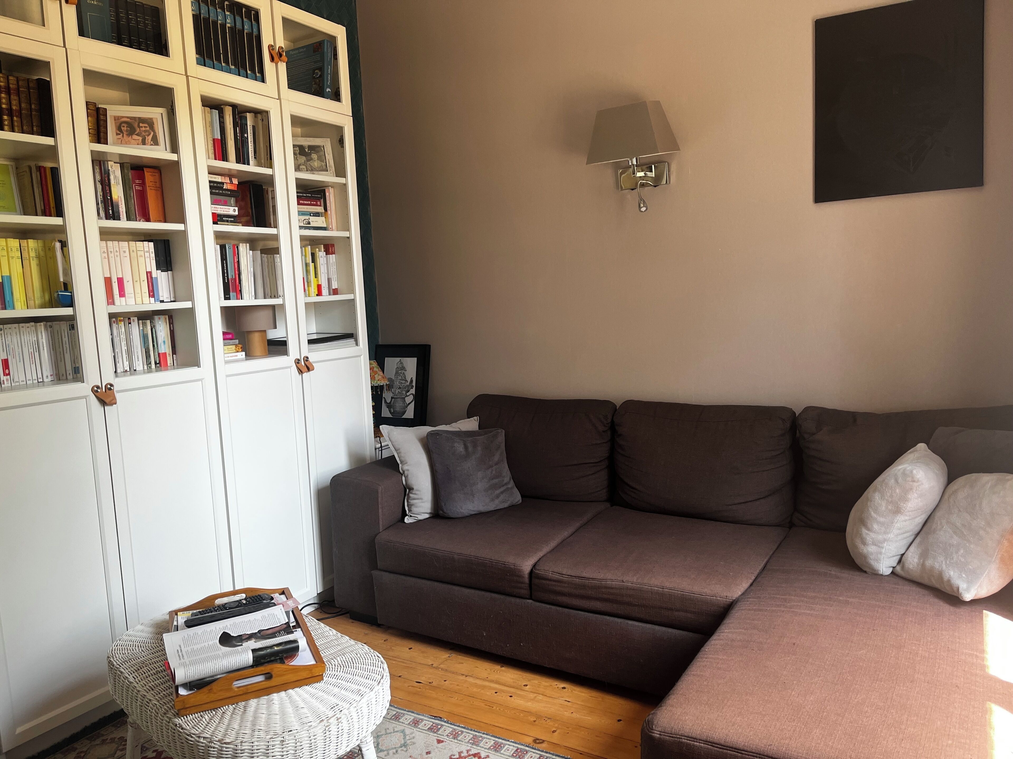 A living room with a brown sofa, a bookshelf, a lamp, and a small round table with books and remote controls.