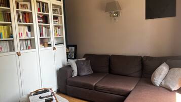A living room with a brown sofa, a bookshelf, a lamp, and a small round table with books and remote controls.