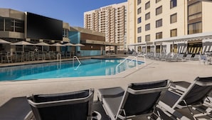Une piscine d’hôtel entourée de chaises longues et de parasols, un grand bâtiment en arrière-plan et un ciel bleu dégagé.