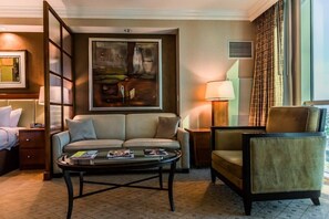 Ein Hotelzimmer mit einer Couch, einem Sessel und einem Couchtisch. An der Wand hängt ein Gemälde und das Fenster ist mit Vorhängen drapiert.