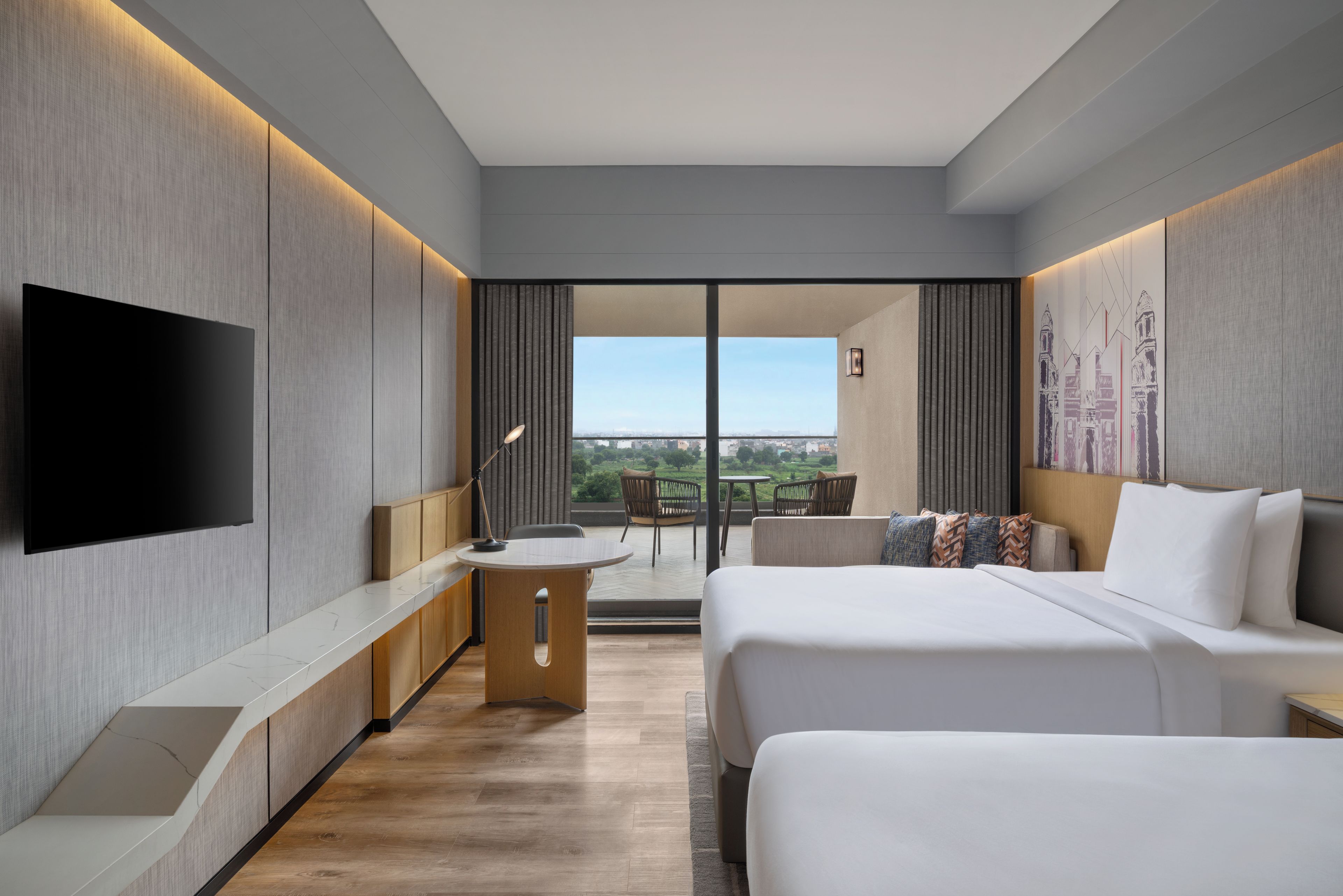 A modern hotel room with a flat-screen TV, a desk with a chair, a bed, a sofa, and a view of the cityscape.