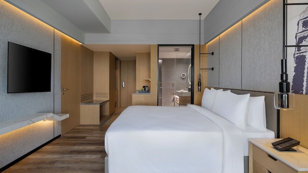 A modern hotel room with a large bed, a flat-screen TV, a desk with a phone, and a bathroom visible through an open door.
