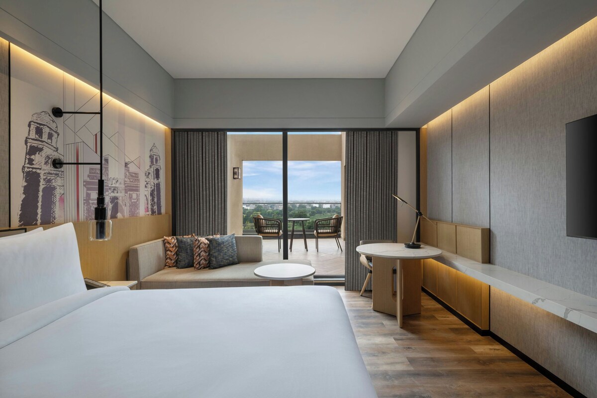 A modern hotel room with a large bed, a sofa, a small table, and a balcony with a view.
