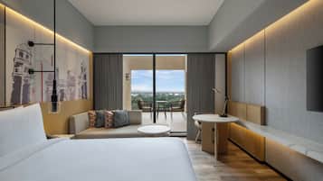 A modern hotel room with a large bed, a sofa, a small table, and a balcony with a view.
