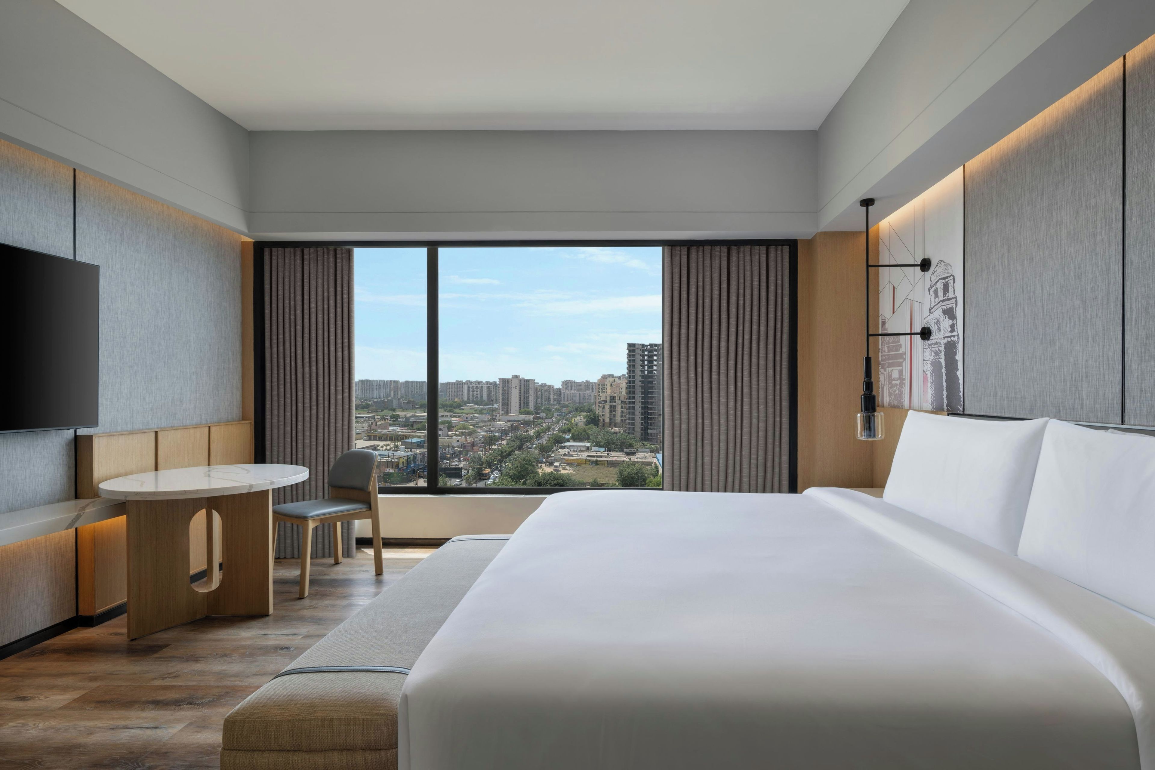 A modern hotel room with a large bed, a desk with a chair, a flat-screen TV, and a city view through the window.