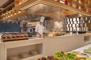 A modern kitchen with a chef preparing food, stainless steel appliances, and a variety of cooking utensils and ingredients. - Courtyard by Marriott Ghaziabad India (Ghaziabad)