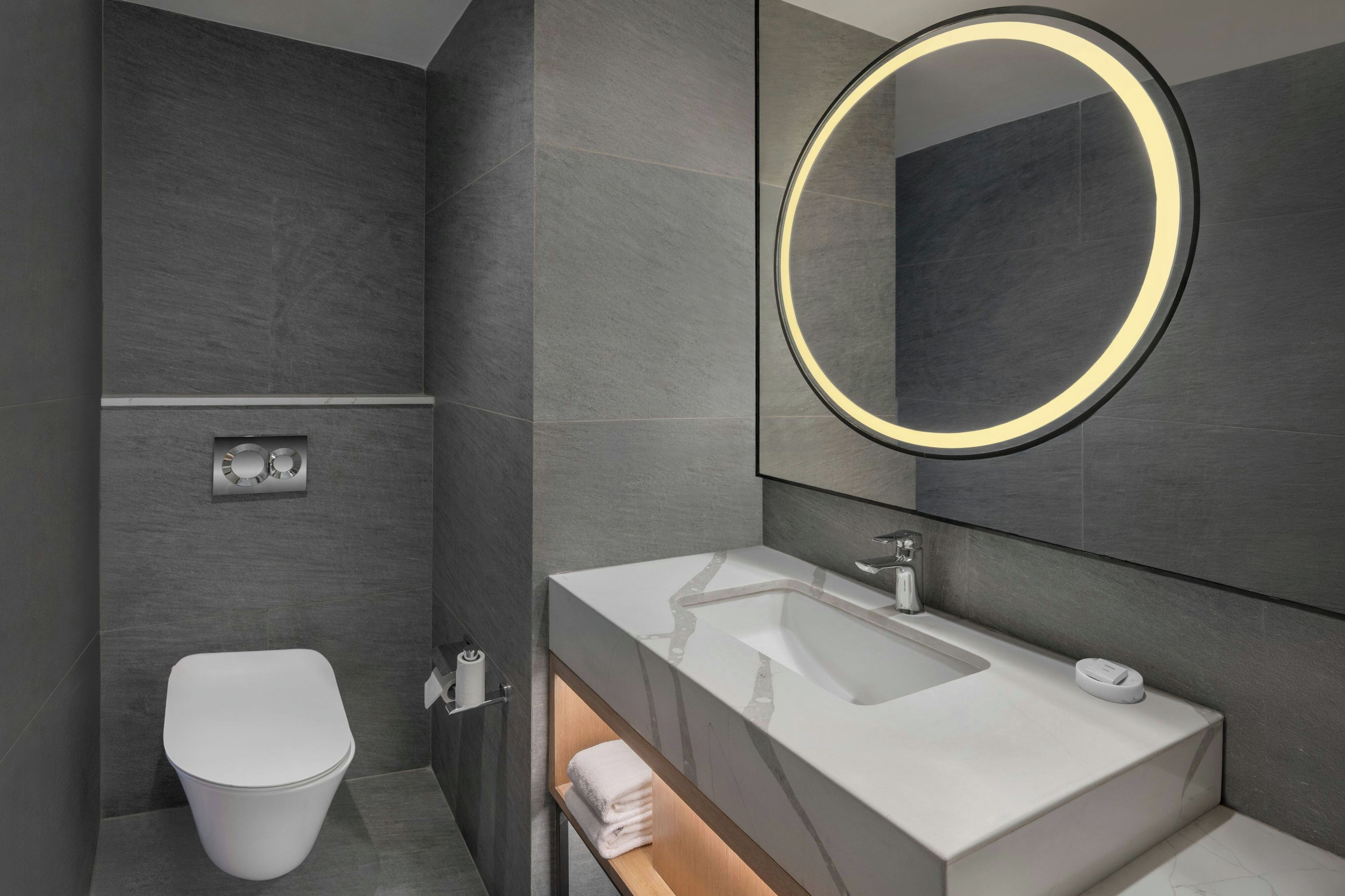 Une salle de bain moderne avec des toilettes blanches, un lavabo doté d’un plan de travail en marbre et un miroir avec un éclairage circulaire.