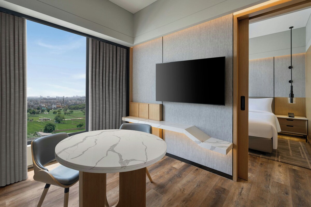 A modern hotel room with a large window, a flat-screen TV mounted on the wall, a round marble table, and a bed with a wooden headboard.