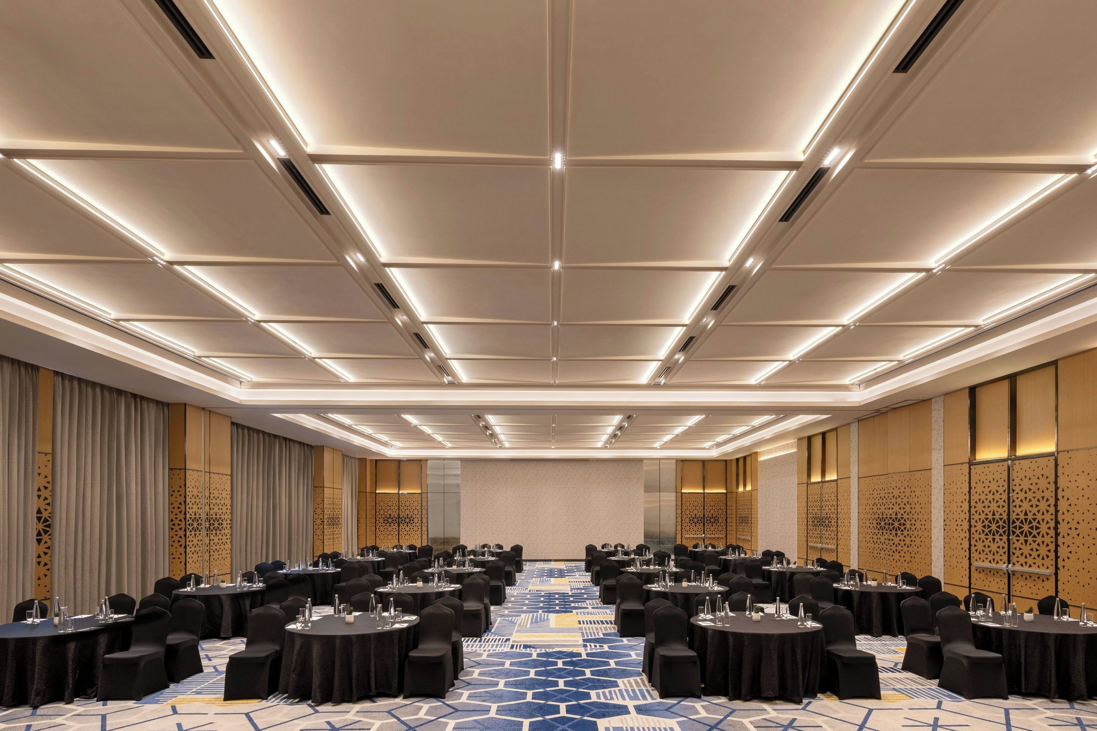 Un salón de conferencias con varias mesas y sillas, una alfombra estampada e iluminación empotrada en el techo.