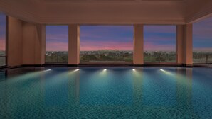 A large indoor swimming pool with clear blue water, surrounded by walls and large windows offering a view of a cityscape at dusk. - Courtyard by Marriott Ghaziabad India (Ghaziabad)