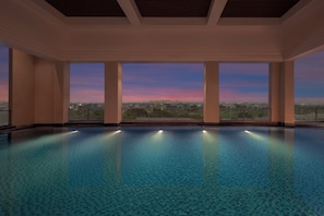 A large indoor swimming pool with clear blue water, surrounded by walls and large windows offering a view of a cityscape at dusk. - Courtyard by Marriott Ghaziabad India (Ghaziabad)