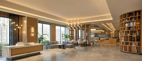 A modern hotel lobby with a reception desk, seating area, and a kitchen visible in the background.