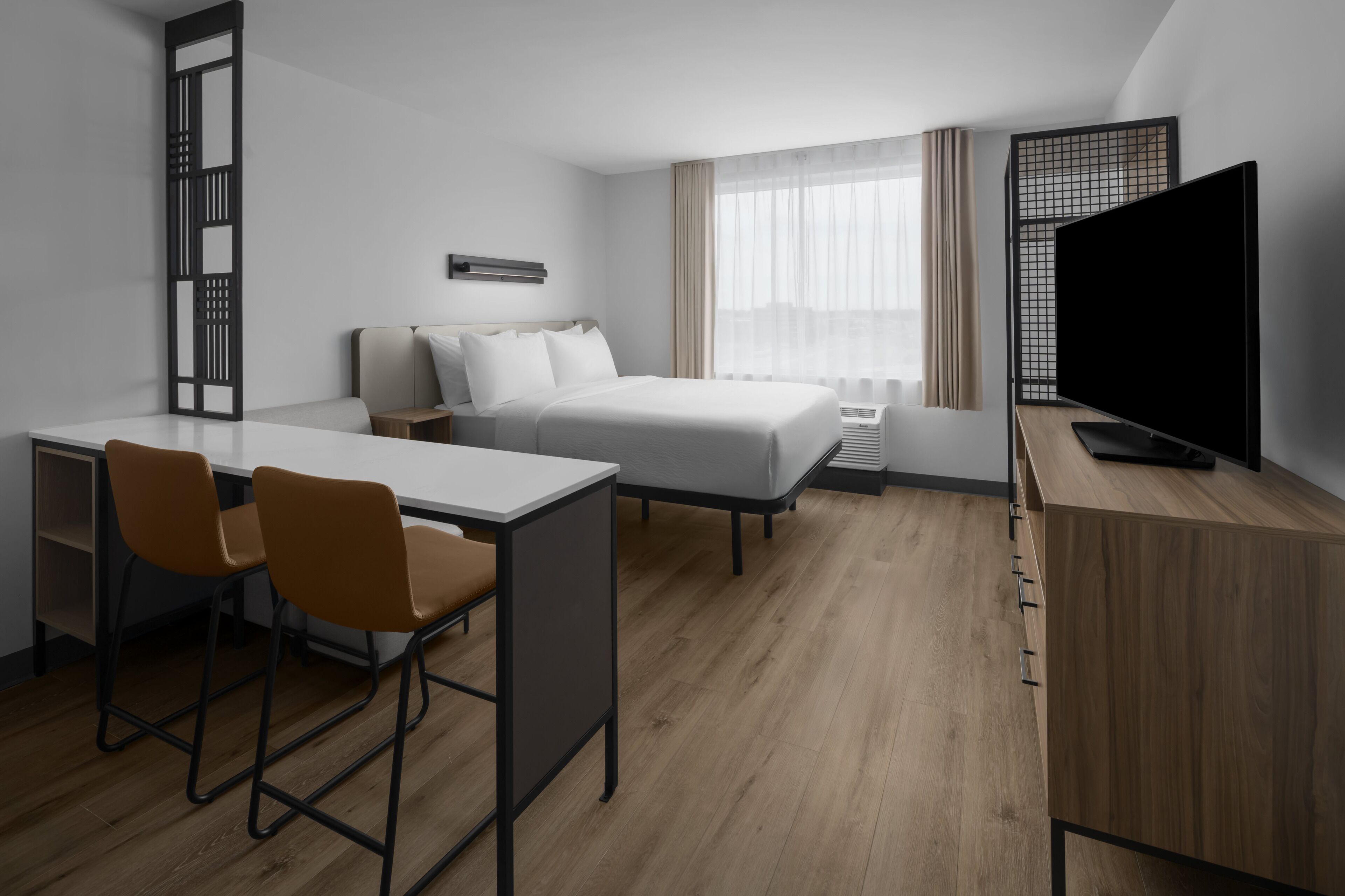 A hotel room with a bed, a desk with two chairs, a television, and a window with curtains.