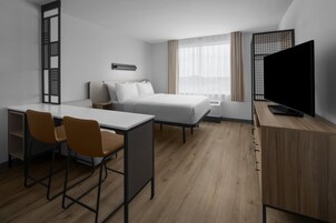 Una habitación de hotel con cama, escritorio con dos sillas, televisor y ventana con cortinas.