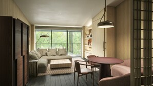 Un interior moderno con un sofá, una mesa de centro, sillas, una estantería de madera y un ventanal con vistas a árboles.