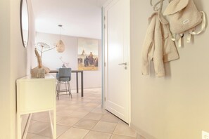 Un pasillo con una puerta blanca, un comedor con mesa y sillas, y un perchero con un abrigo color beige.