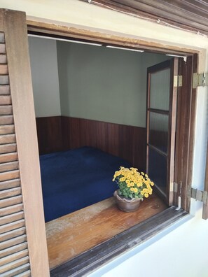 A room with a wooden-framed window, a blue bed, and a potted plant with yellow flowers.