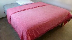 Un llit individual amb llençol rosa i almoina blanca en una habitació amb paret senzilla i una petita prestatgeria de fusta.