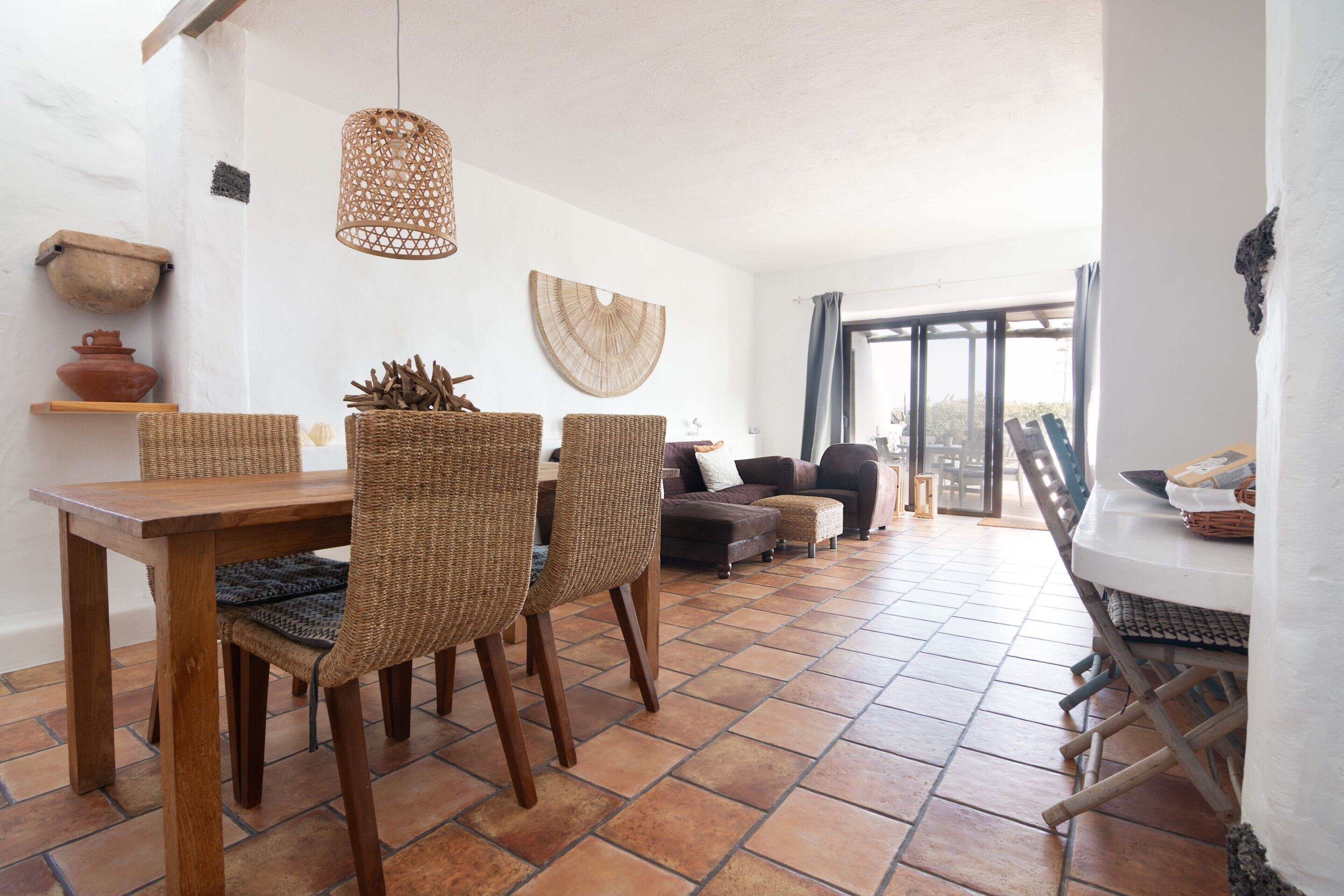 Uma área de refeições com mesa de madeira e cadeiras de fibra natural, luminária pendente trançada e vista para um pátio externo através de uma porta de vidro.