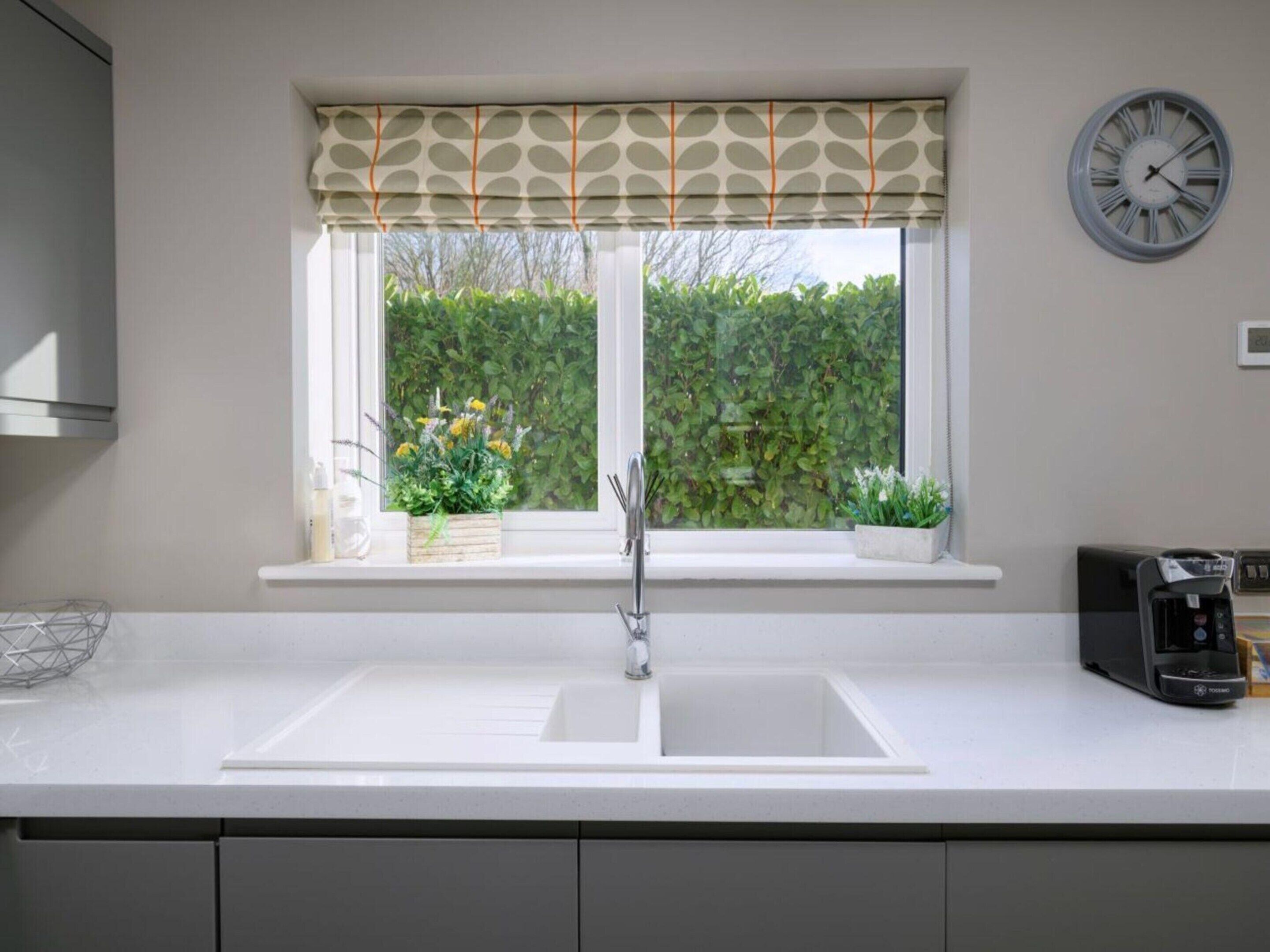 A kitchen with a white countertop, a sink, a window with a patterned valance, and a wall-mounted clock.