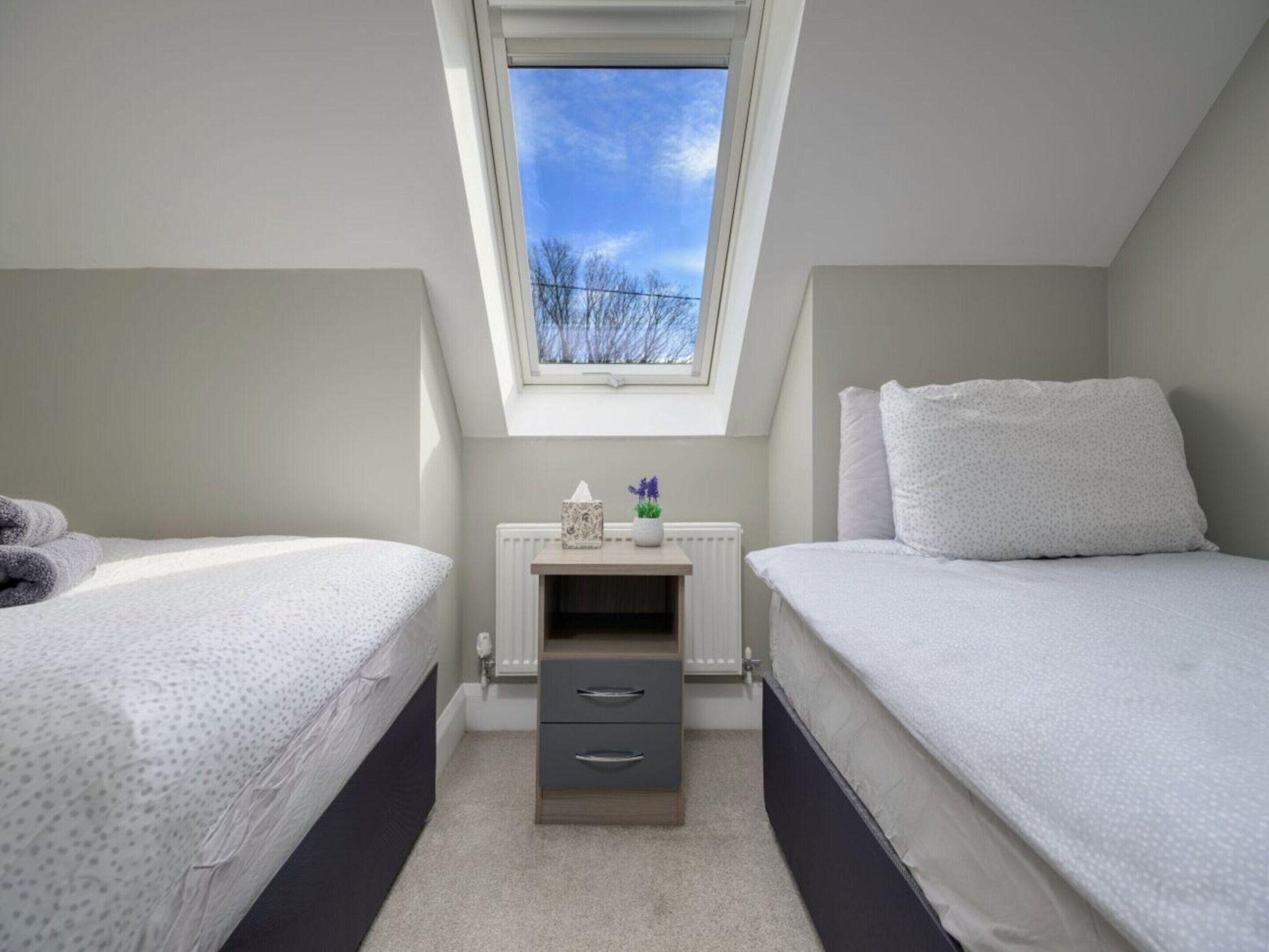A bedroom with two beds, a small bedside table with drawers, a radiator, and a window with a view of trees.