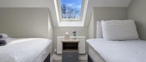 A bedroom with two beds, a small bedside table with drawers, a radiator, and a window with a view of trees.