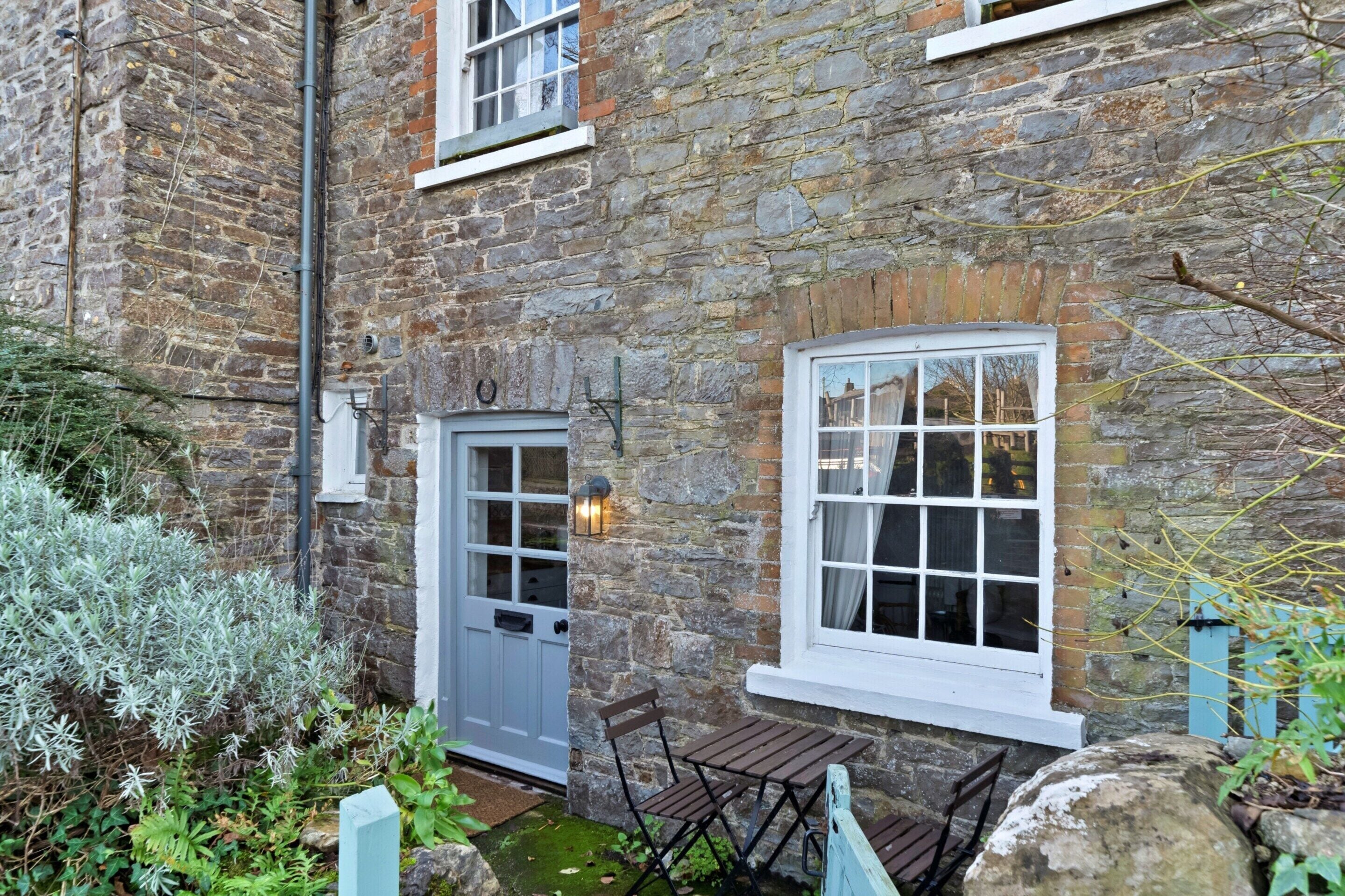 A stone building with a white door, a window with a light fixture, and a small outdoor seating area with a bench and table.