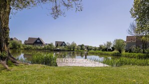 A serene lakeside setting with a wooden dock extending into the water, surrounded by lush greenery and residential buildings. - Compact Waterfront Wellness Bungalow (Makkum)