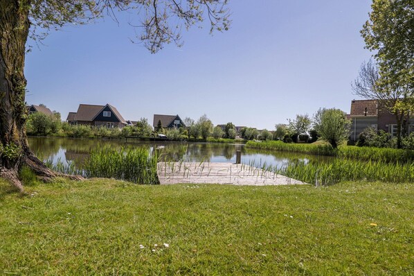 A serene lakeside setting with a wooden dock extending into the water, surrounded by lush greenery and residential buildings. - Compact Waterfront Wellness Bungalow (Makkum)
