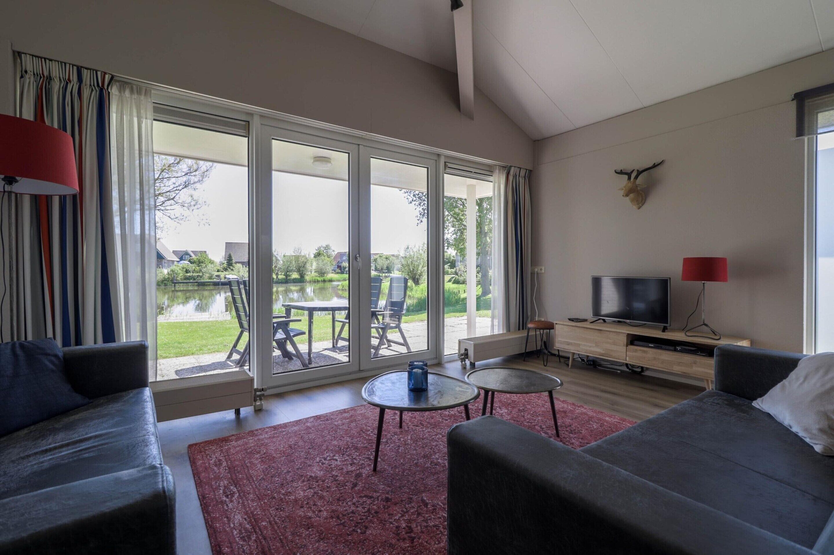 A modern living room with a large sliding glass door leading to a patio area. There is a television on a wooden stand, a red lamp, and a mounted deer head on the wall.