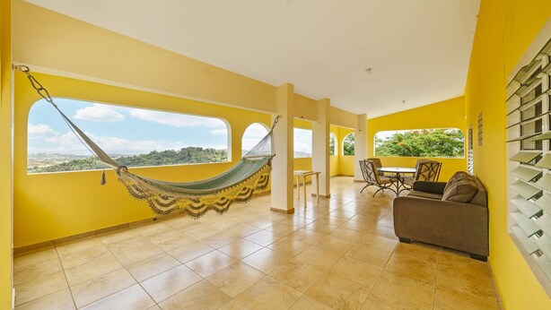 A spacious room with yellow walls, a hammock, a sofa, and a dining area with a view. - Hilltop Getaway With Huge Terrace (Fajardo)