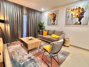 Uma sala de estar moderna com um sofá, mesa de centro e uma poltrona. Há duas pinturas na parede e uma planta no canto.