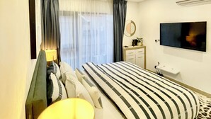 A modern hotel room with a large bed, a bedside table with a lamp, a flat-screen TV mounted on the wall, and a window with curtains. - Stylish East Legon 1BR | Pool & Gym (Accra)