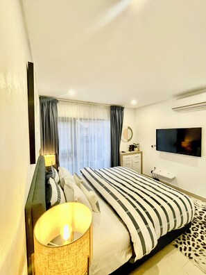 Quarto de hotel moderno com uma cama grande, uma mesa de cabeceira com abajur, uma TV de tela plana na parede e uma janela com cortinas.