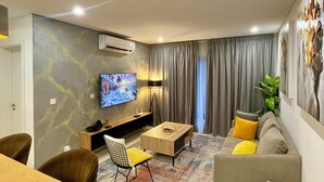 A modern living room with a flat-screen TV, a wooden coffee table, a sofa with cushions, and a dining area with a table and chairs. - Stylish East Legon 1BR | Pool & Gym (Accra)