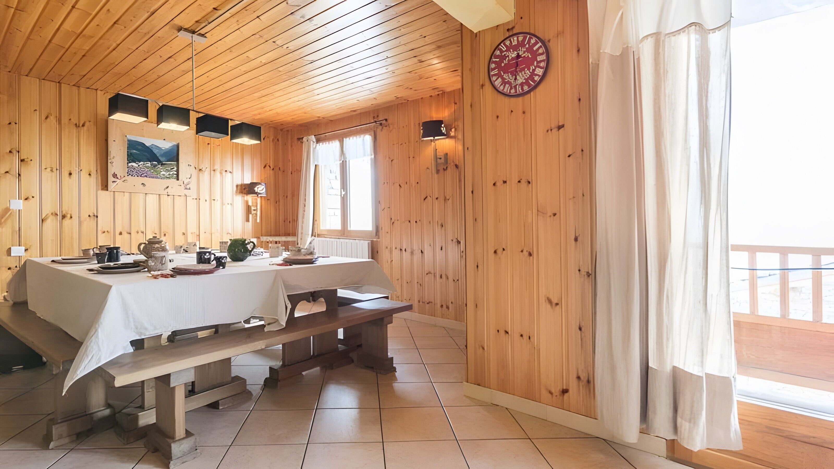 Interior de madera con una mesa de comedor lista para cuatro personas, un banco y un reloj en la pared.
