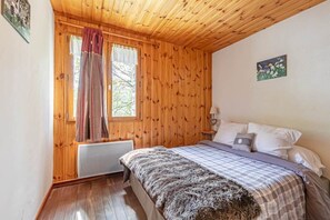 Ein Schlafzimmer mit Holzwänden, einem Bett mit kariertem Überwurf, einem Fenster mit Vorhängen und einem kleinen Nachttisch mit Lampe.