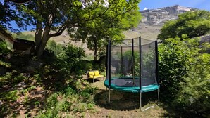 Un trampolín con red de seguridad, rodeado de follaje y una silla amarilla, con un paisaje montañoso al fondo.