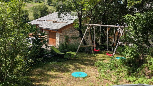 Una casa de piedra con techo de tejas, un columpio y una tapa de alcantarilla azul en un jardín.